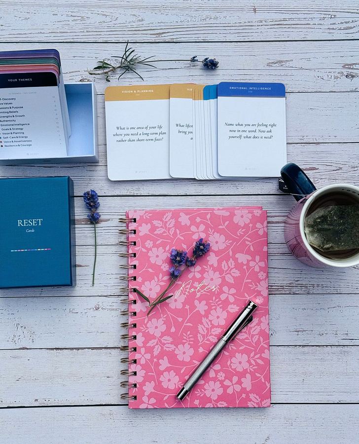 The RESET Cards deck fanned open beside its box, a notebook and a cup of tea on a wooden table