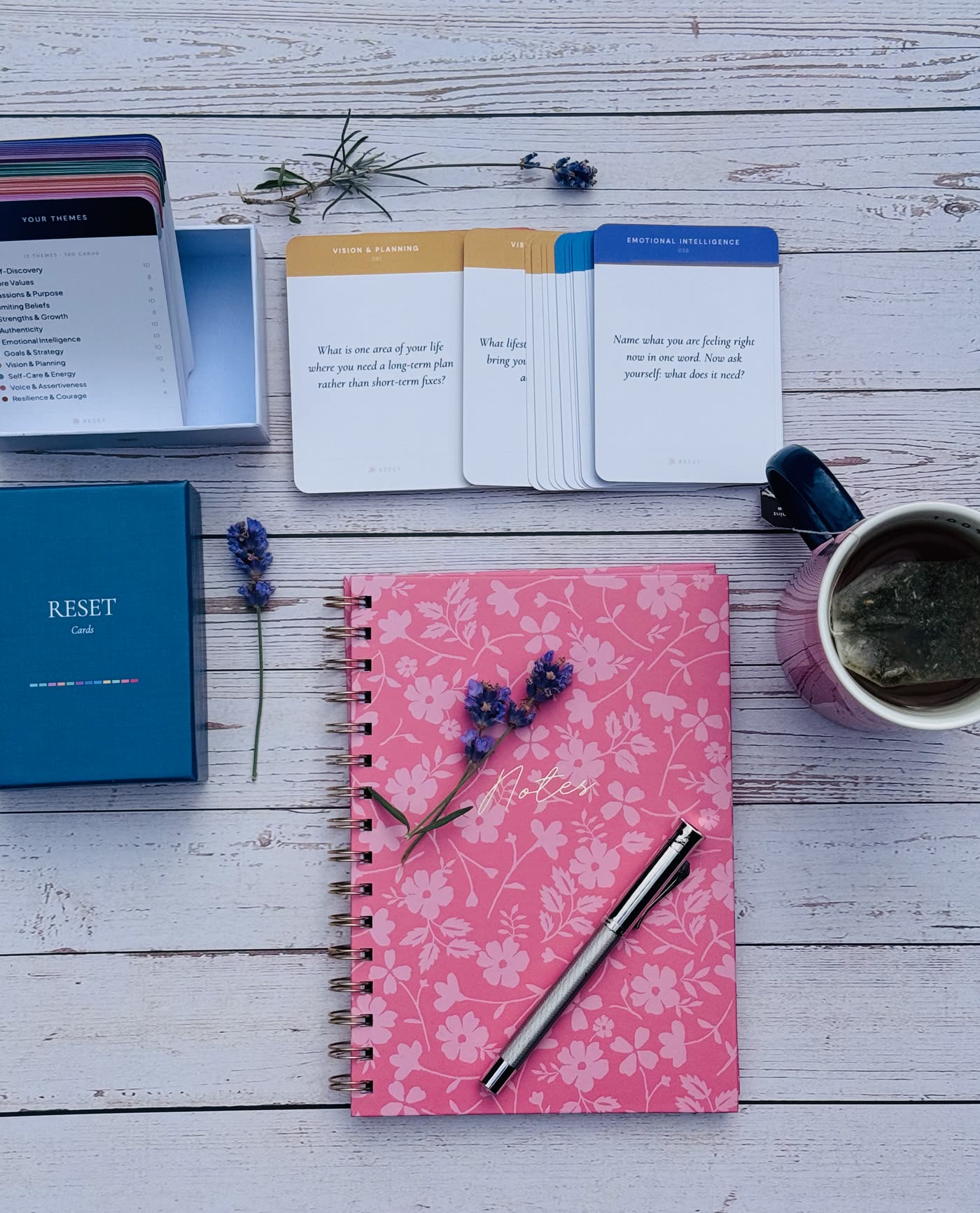 The RESET deck open on a white wooden table with drawn cards, a pink notebook, lavender and a cup of tea